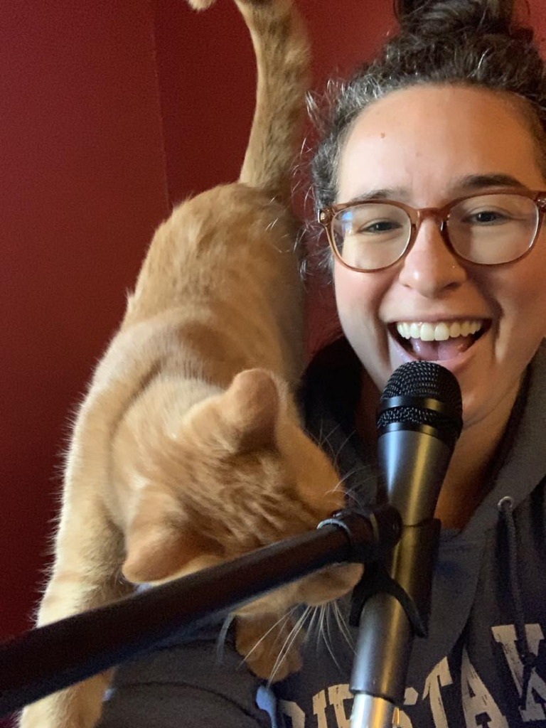 Rachel Mousie speaking into microphone, smiling, with cat cozying up next to her.