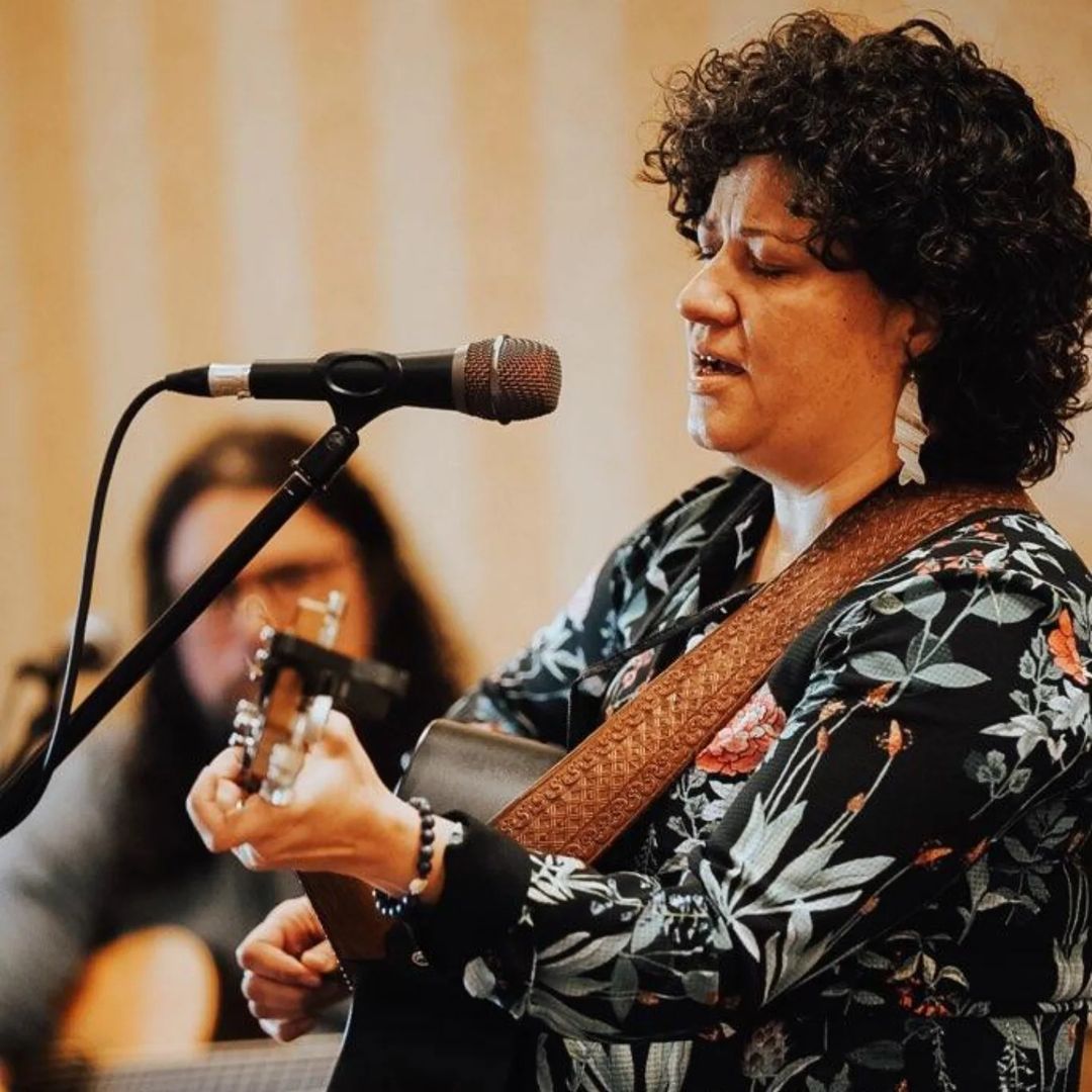 Elexa Dawson sings into microphone, playing guitar, while Aaron looks on.