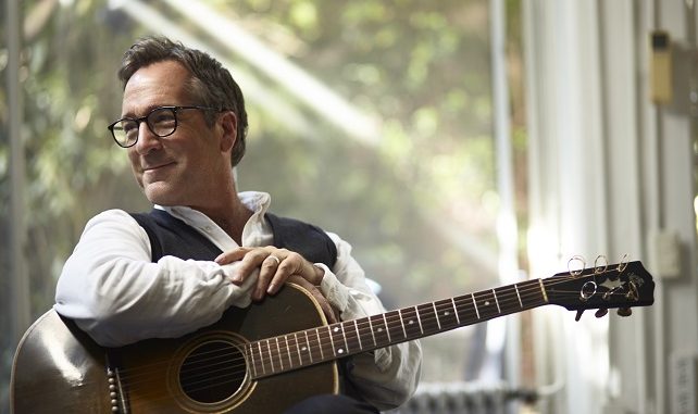 Richard Shindell looks out a window, smiling and holding a guitar.