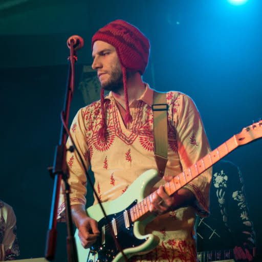 Burr Settles at a club, wearing a winter hat and holding an electric guitar, bathed in blue light.