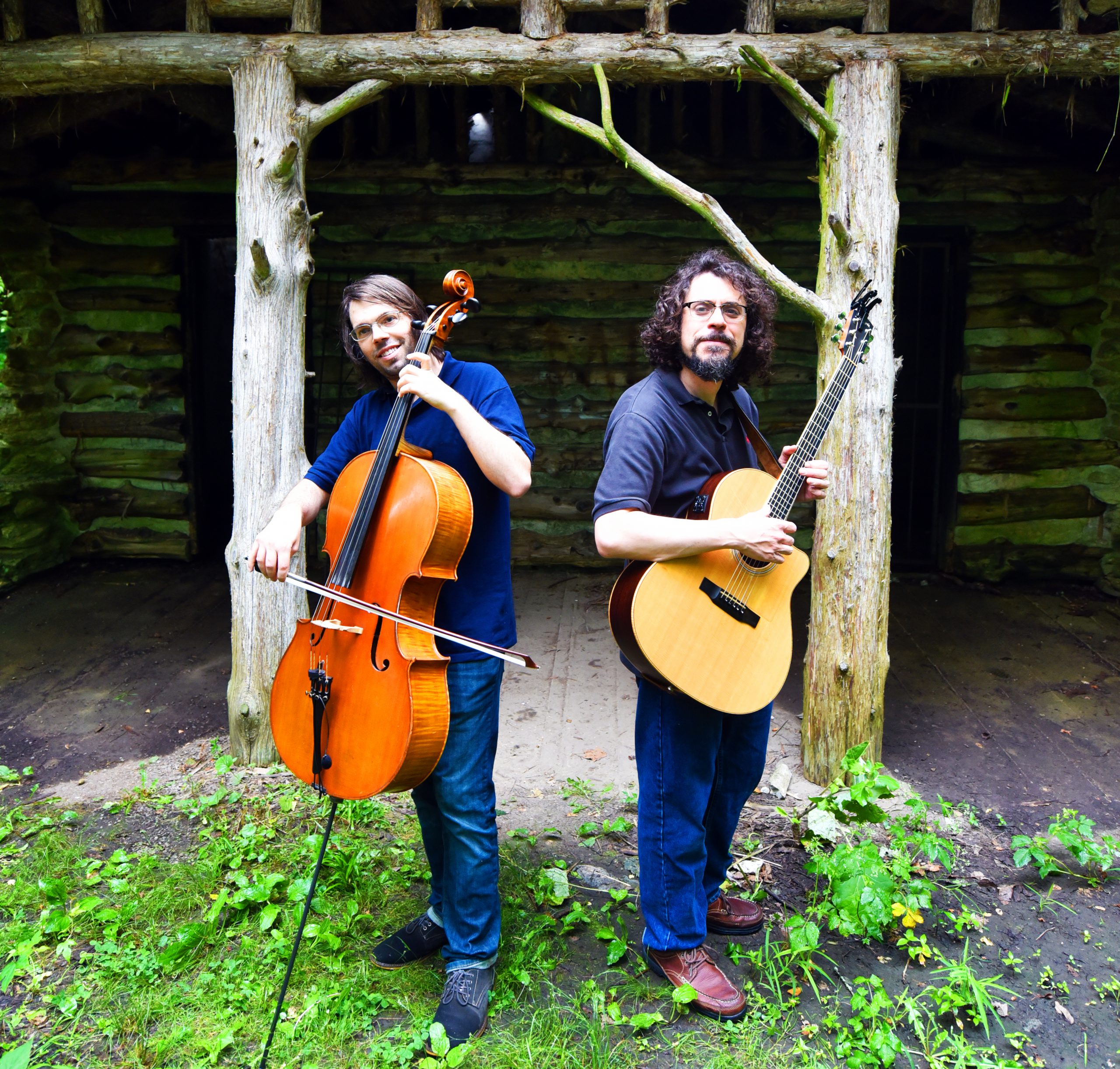 Aaron and Michael hold their instruments in front of a wooden hut.