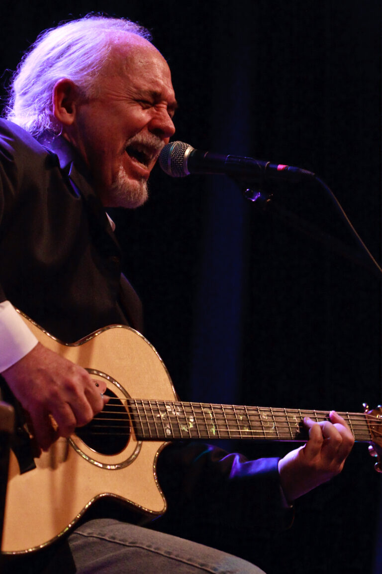 Craig Bickhardt sings into microphone and strums guitar. Dark background.